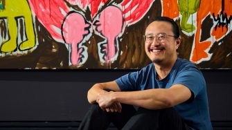 A smiling man sitting in front of a colourful mural with his arms crossed.