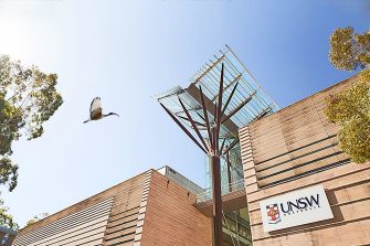 Looking up at the top of  Scientia building with the glass overhang cover, trees at each side and an ibis flying past in clear blue sky.