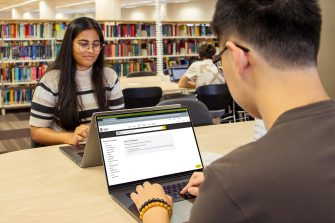 Two students inside the Library, sitting opposite each other at a large desk and using their own laptops.