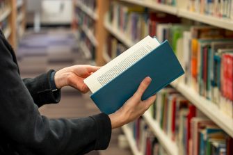 Borrowing A person standing amongst the shelves in the Main Library, a close up focus of their hands which is flipping through a blue hardcover library book.