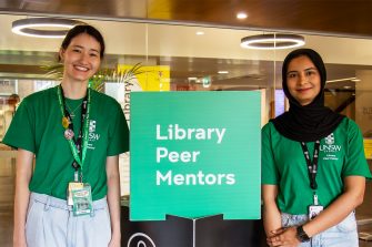 Two Library Peer Mentors standing next to the Library Peer Mentors sign in the Main Library foyer. They are in green uniforms and smiling at the camera.