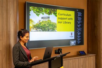 A librarian delivering a workshop presentation, the screen behind her shows a slide with a heading that reads 'Curriculum support with our Library'.