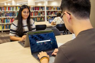 Two students inside the Library, sitting opposite each other at a large desk and using their own laptops. The MedicineComplete database is visible on one of their screens.