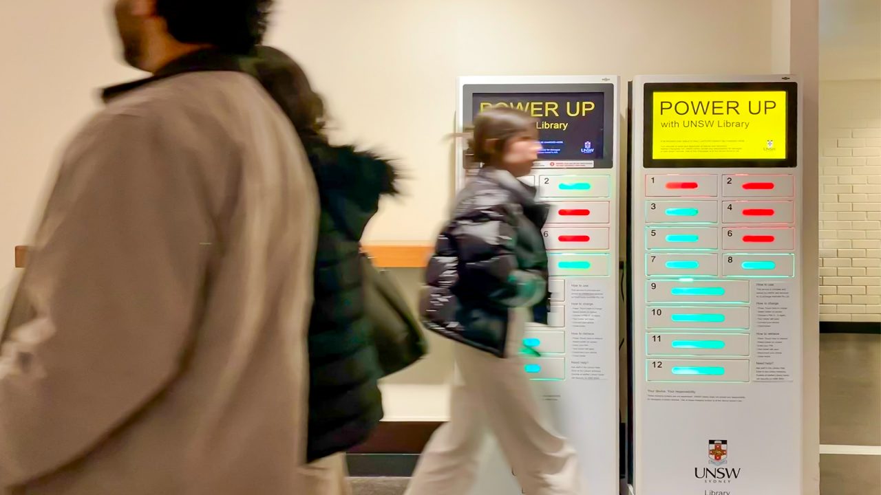 Time lapse showing people walking past two device charging locker stands. "POWER UP with UNSW Library" text with yellow background clear on one of the display screen.