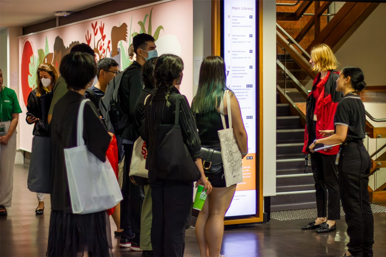 A group of students looking towards the digital floor wayfinder sign at the bottom of the stairwell. Painted wall mural in background.