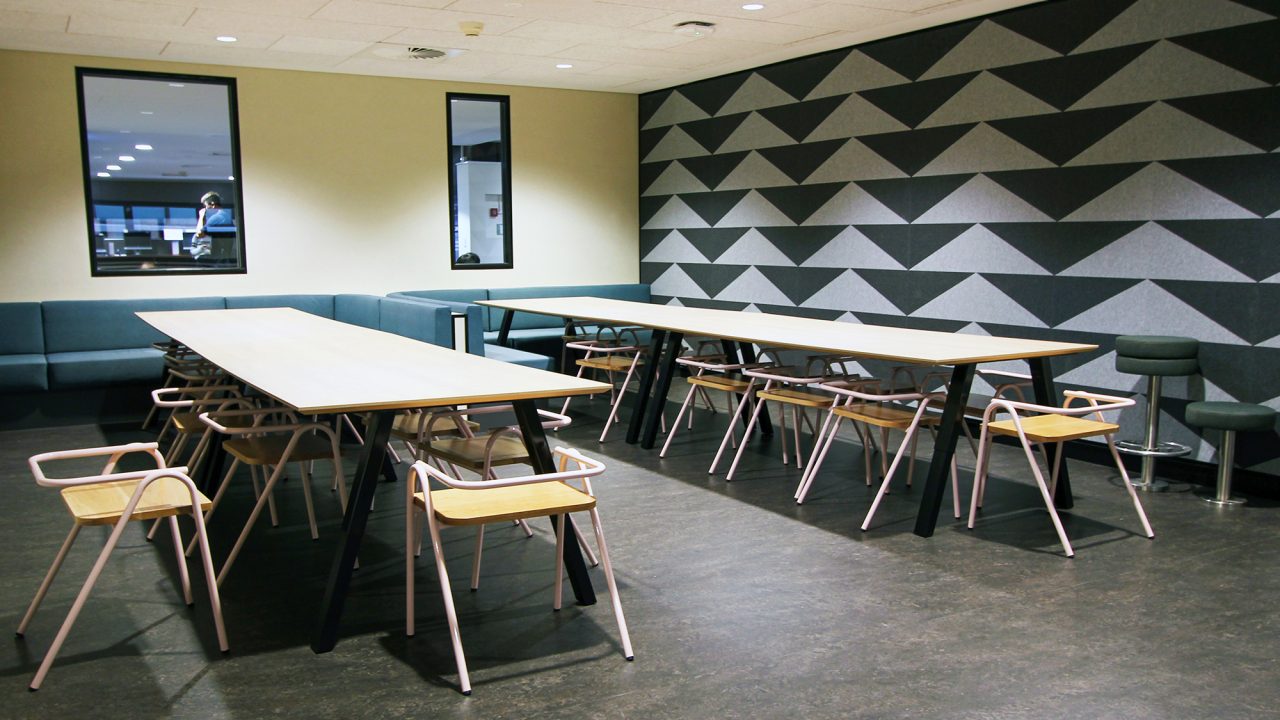 two long bench tables with very low back charis. Blue lounge seats along the rear walls. Abstract triange shapes in dark colours on one wall.
