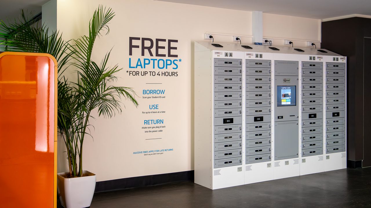 Laptop loan lockers against wall. Black and blue lettering on wall inlcudes "FREE LAPTOPS FOR UP TO 4 HOURS". Palm tree sits to the left side.