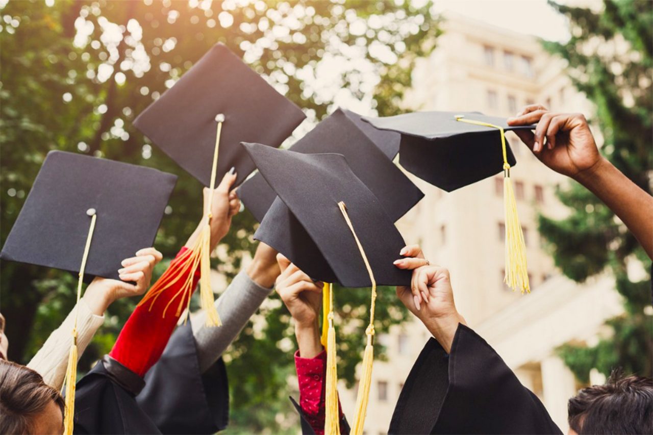 6 arms high in the air, most with black graduation gown sleeves showing, hold black graduation mortar board hats. Trees and building in the distant background.