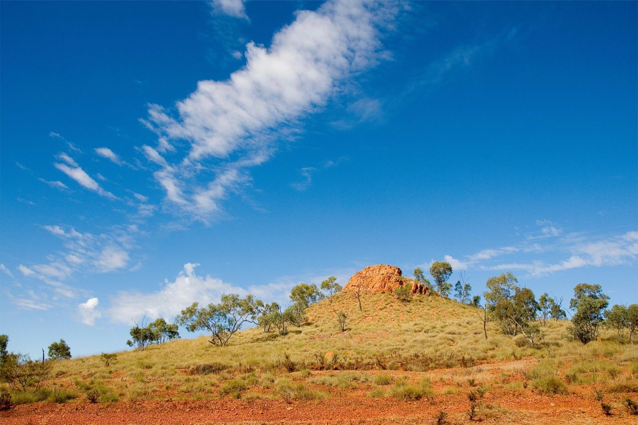 A vast, semi-arid landscape revealing orange dirt, green shrubbery, and a small hill peaking with a cluster of large rocks. 