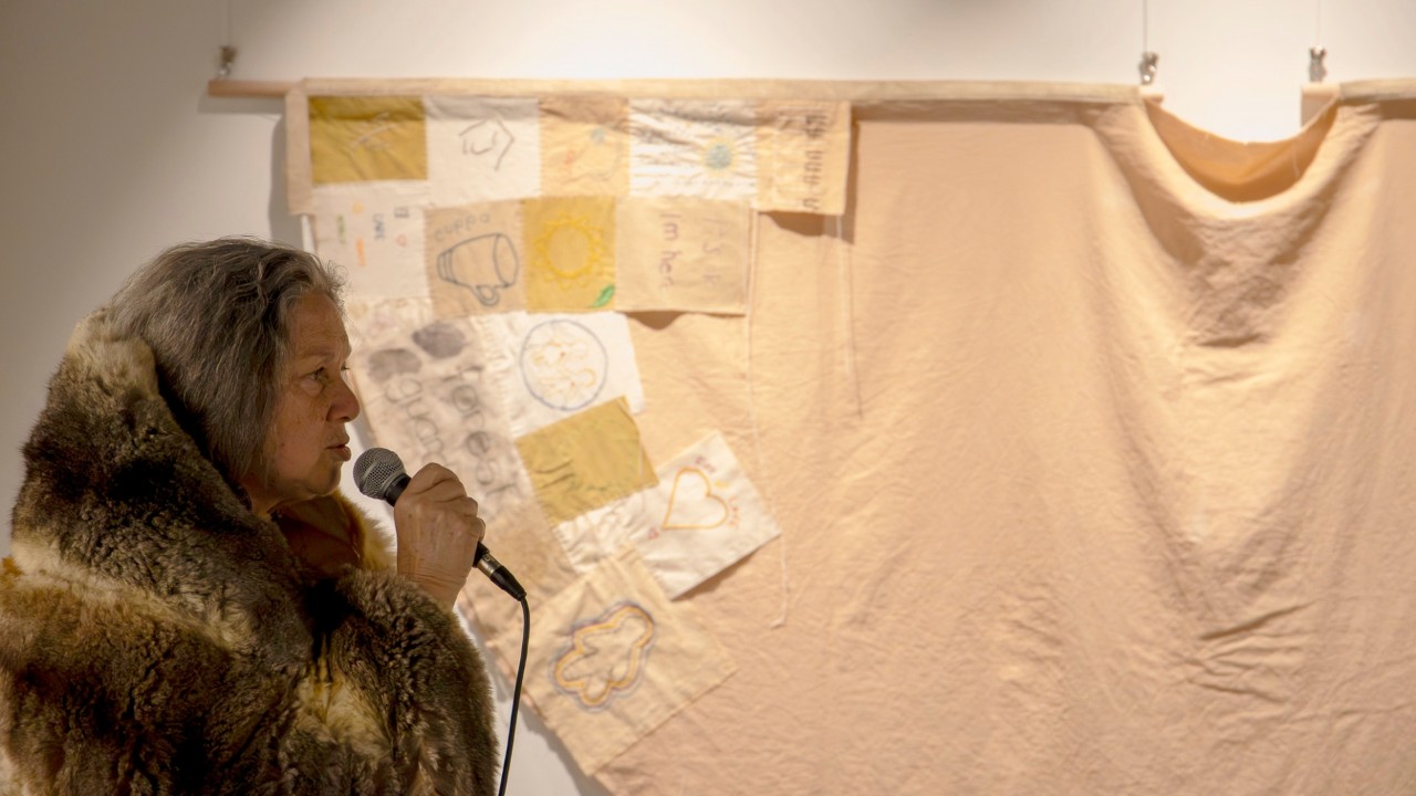 A woman wrapped in pelts speaks into a microphone. Behind her hangs a large dusty pink cloak with embroidered squares.