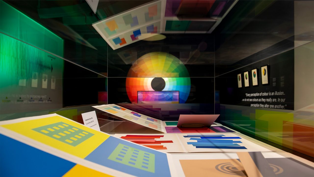 A glass display case filled with images of colourful patterns. In background is a black wall with a large colour-wheel. 