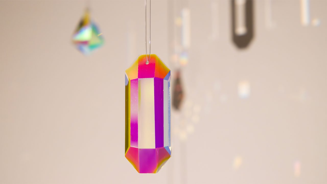 A crystal prism hangs suspended, radiating a spectrum of iridescent colors in a close-up shot with a blurred, light background.