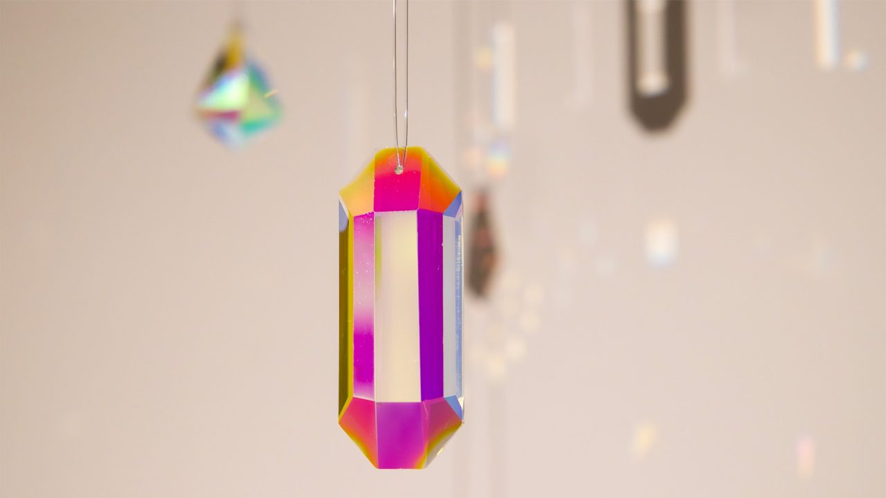 A crystal prism hangs suspended, radiating a spectrum of iridescent colors in a close-up shot with a blurred, light background.