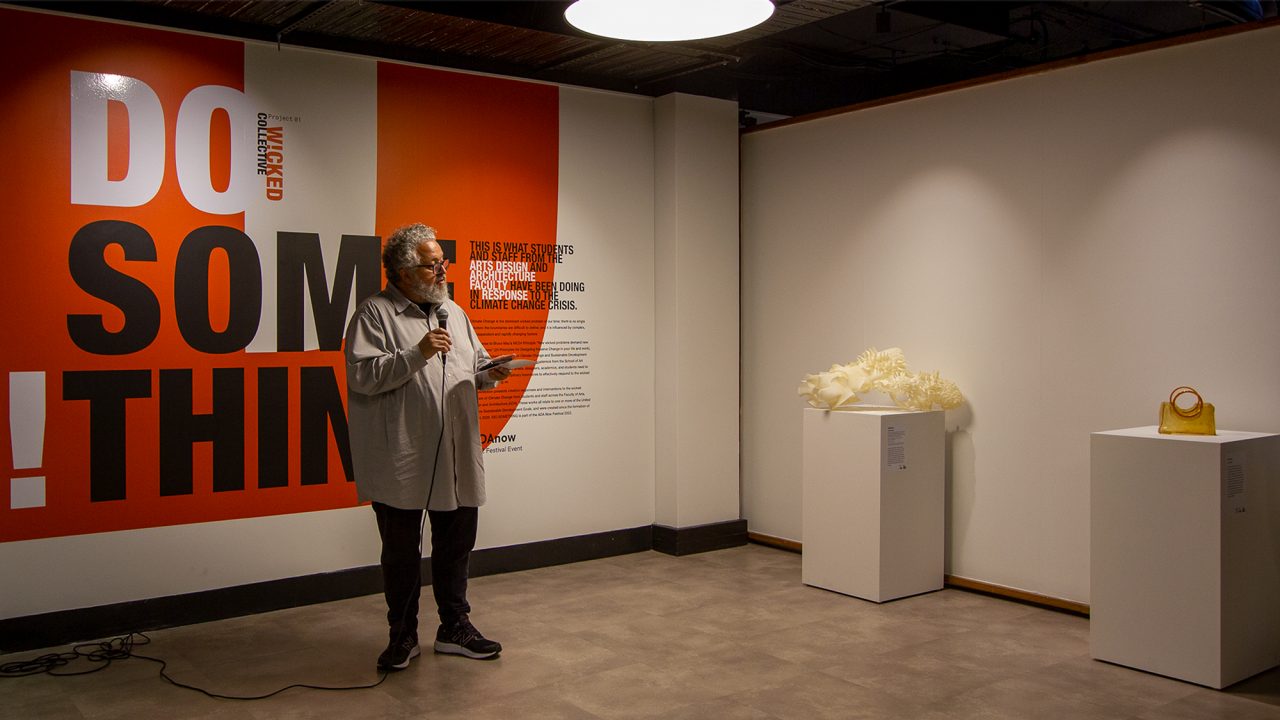 A man holding a microphone delivers a speech in front of an exhibition wall with large text that reads "Do Something".