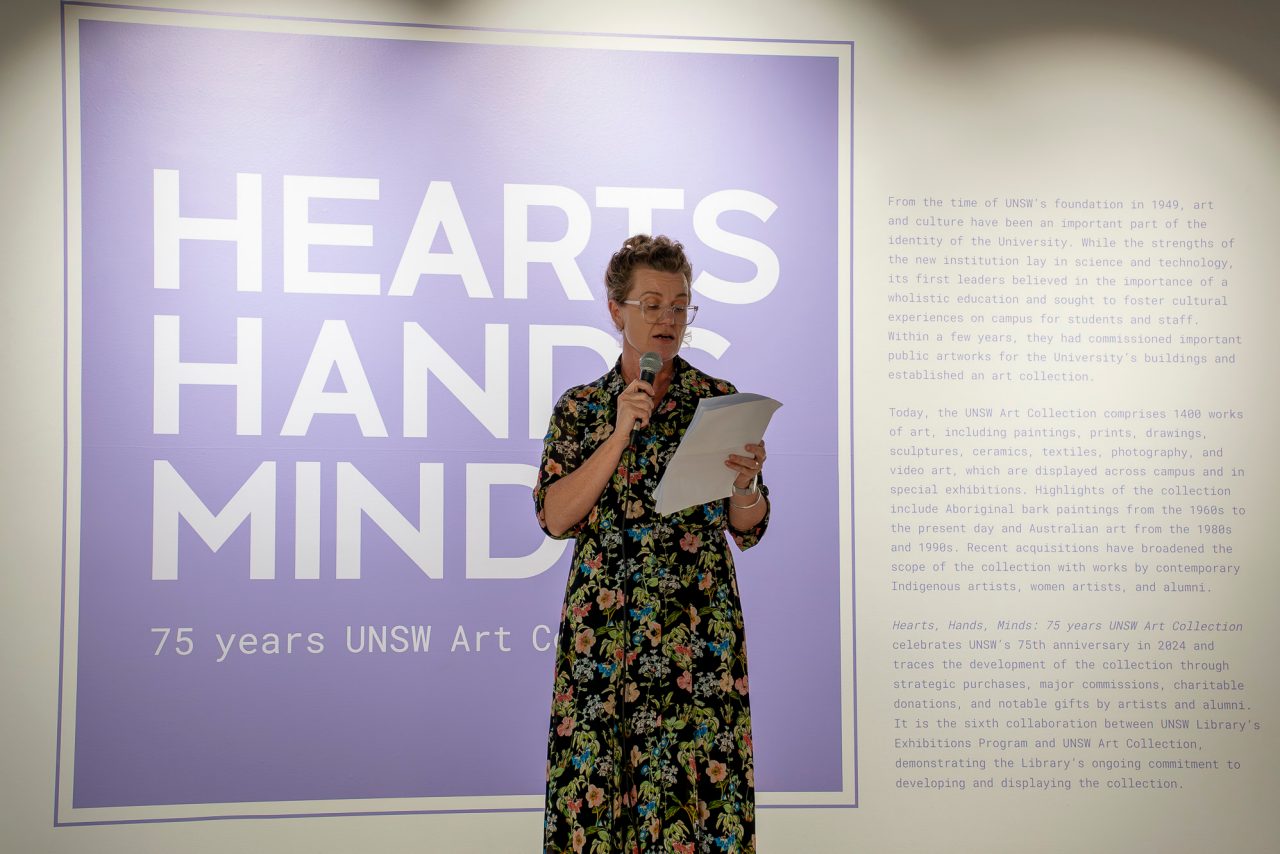 A woman stands in front of a wall with purple text, holding a microphone addressing an audience