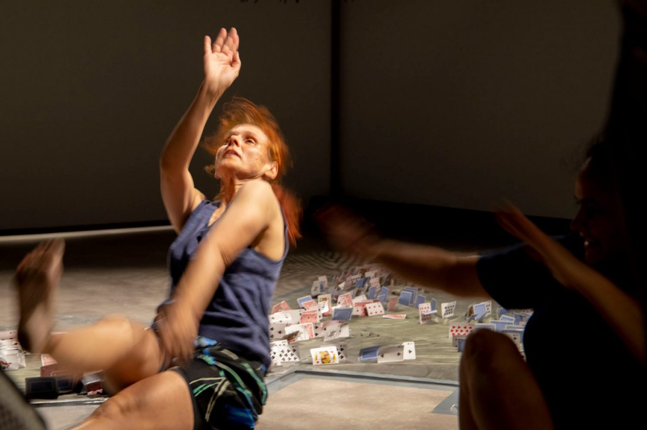 Vicki Van Hout performing seated on floor with playing card installation behind