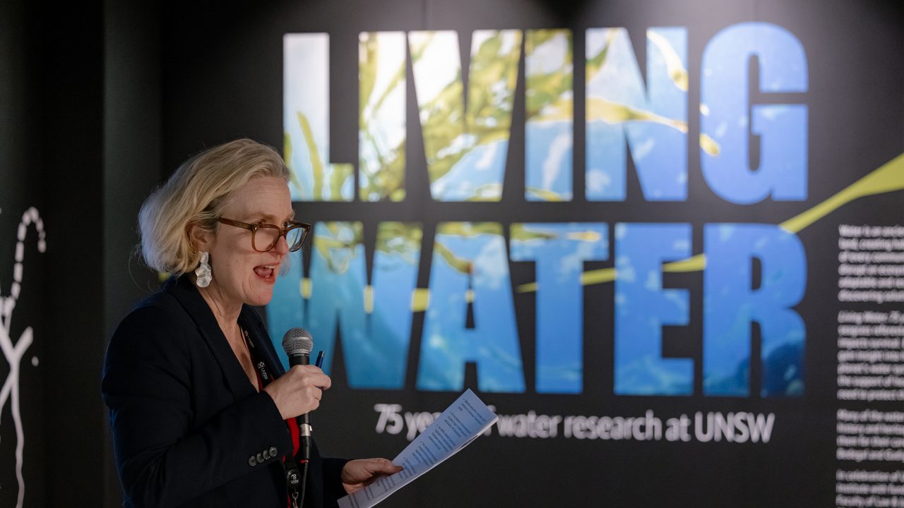 A lady with white hair holds a microphone in one hand in mid-speech. In the background is a wall that says "Living Water".