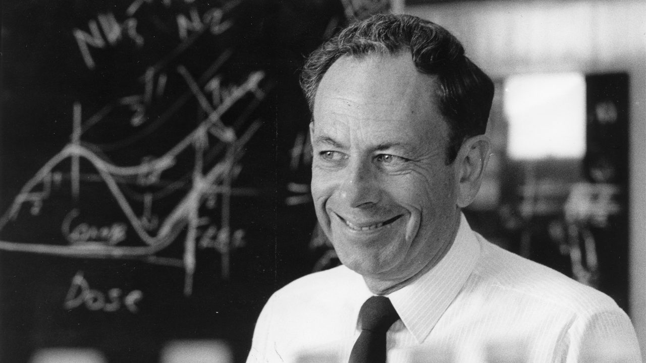 Black and white photo of a smiling man with a blackboard with mathematical equations in the background