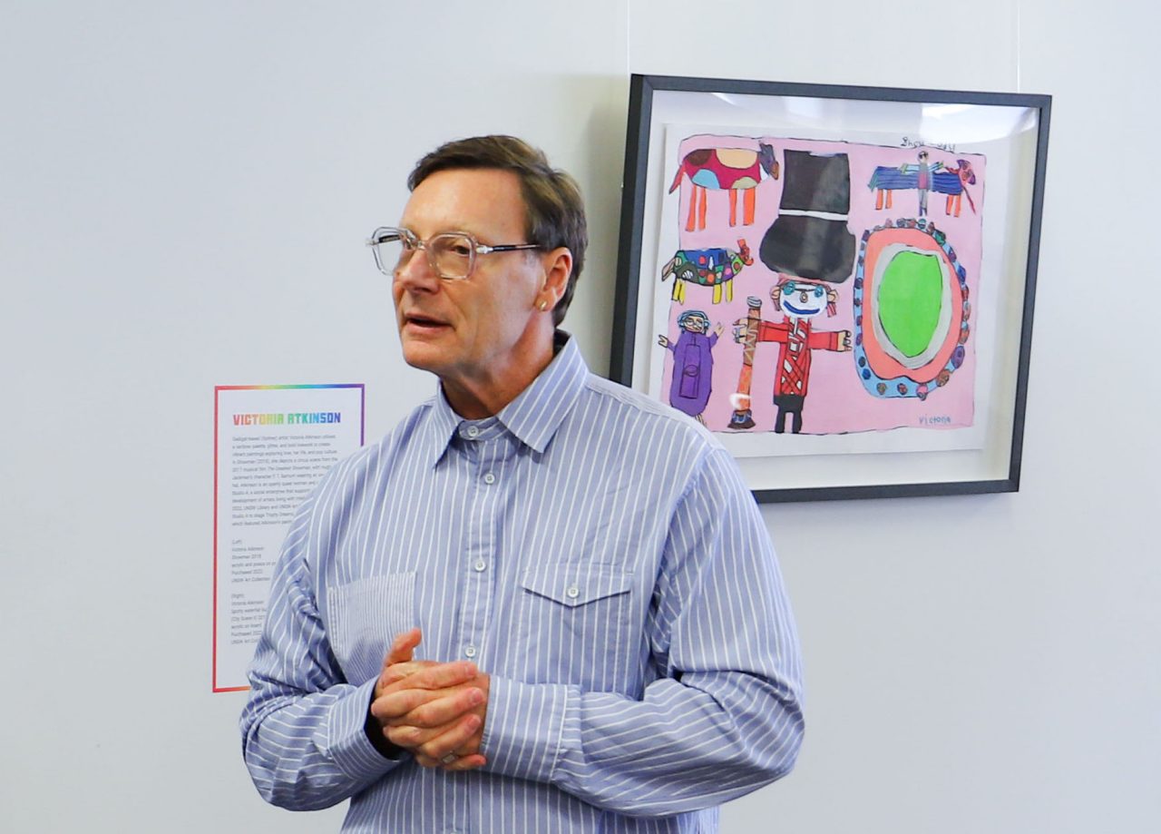 A man in a a blue shirt giving a speech in front of a colourful artwork.