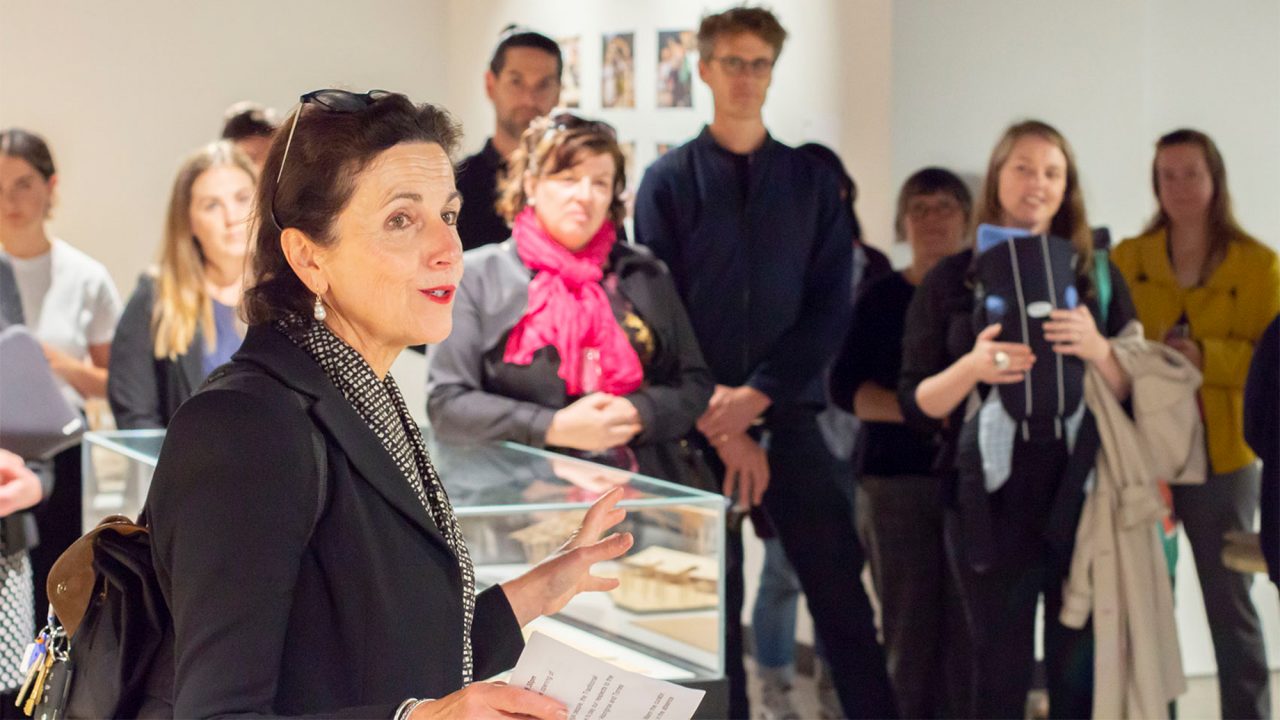 A photo of several people gathered around a display cabinet. A woman wearing a black jacket gestures towards the display with her hand mid-speech.