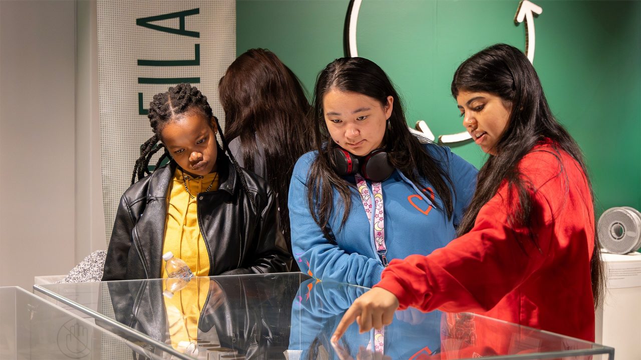 A group of students looking through an exhibition glass cabinet. One of them is pointing to an object inside the cabinet.