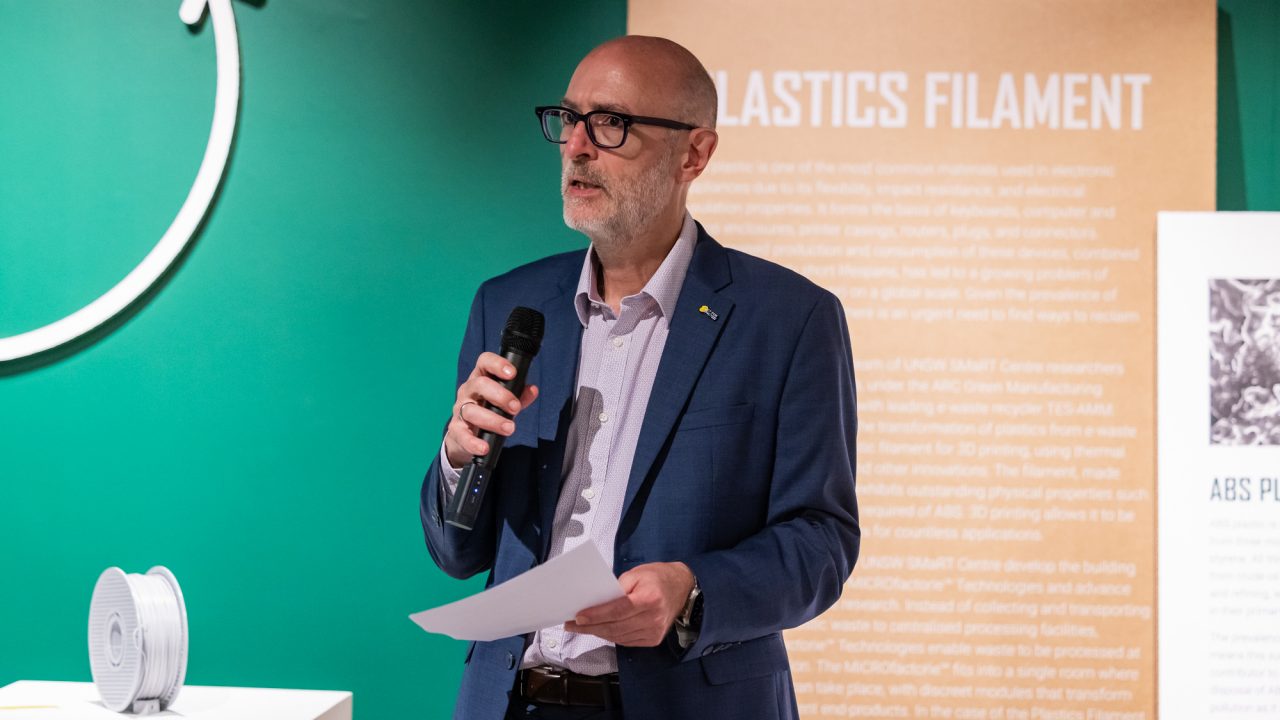 A man holds a microphone in one hand, and a sheet of paper in the other hand, delivering a speech in a gallery space. 