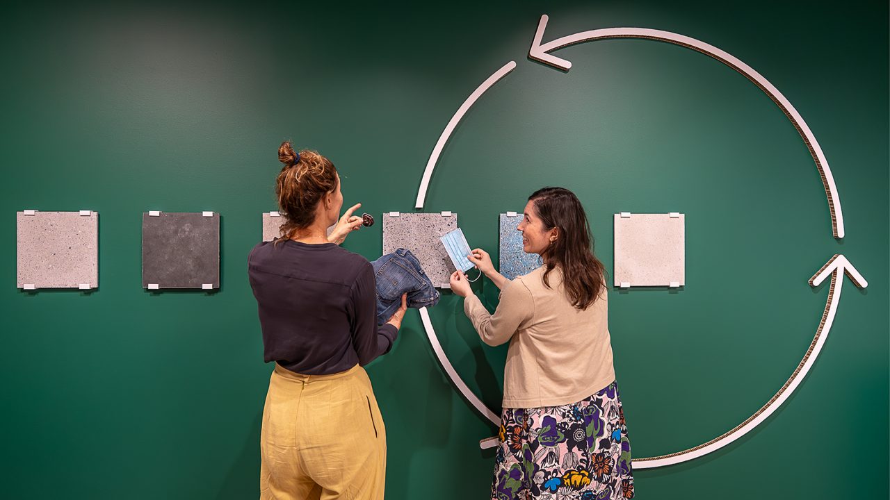 Two women stand into front of a green wall that has six tiles mounted to it. The women hold different types of fabric in their hands, and compare them to the tiles thoughtfully. 