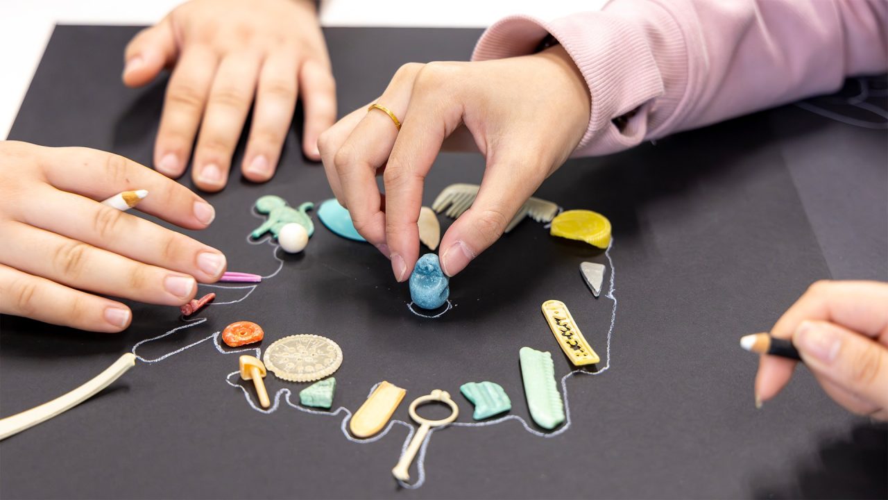 Close up of two people's hands, they are interacting with small colourful objects on the table and drawing a border around the objects.