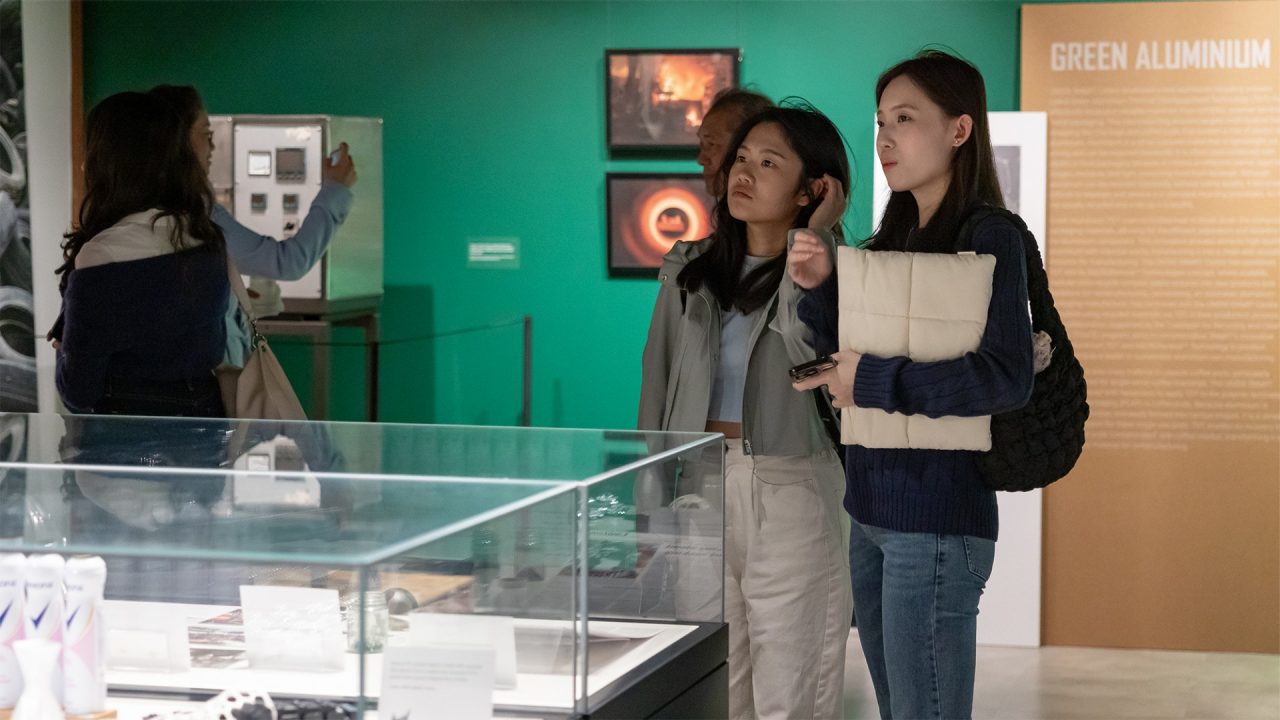 Two women stand in a gallery space with green walls, looking at the exhibits with interest