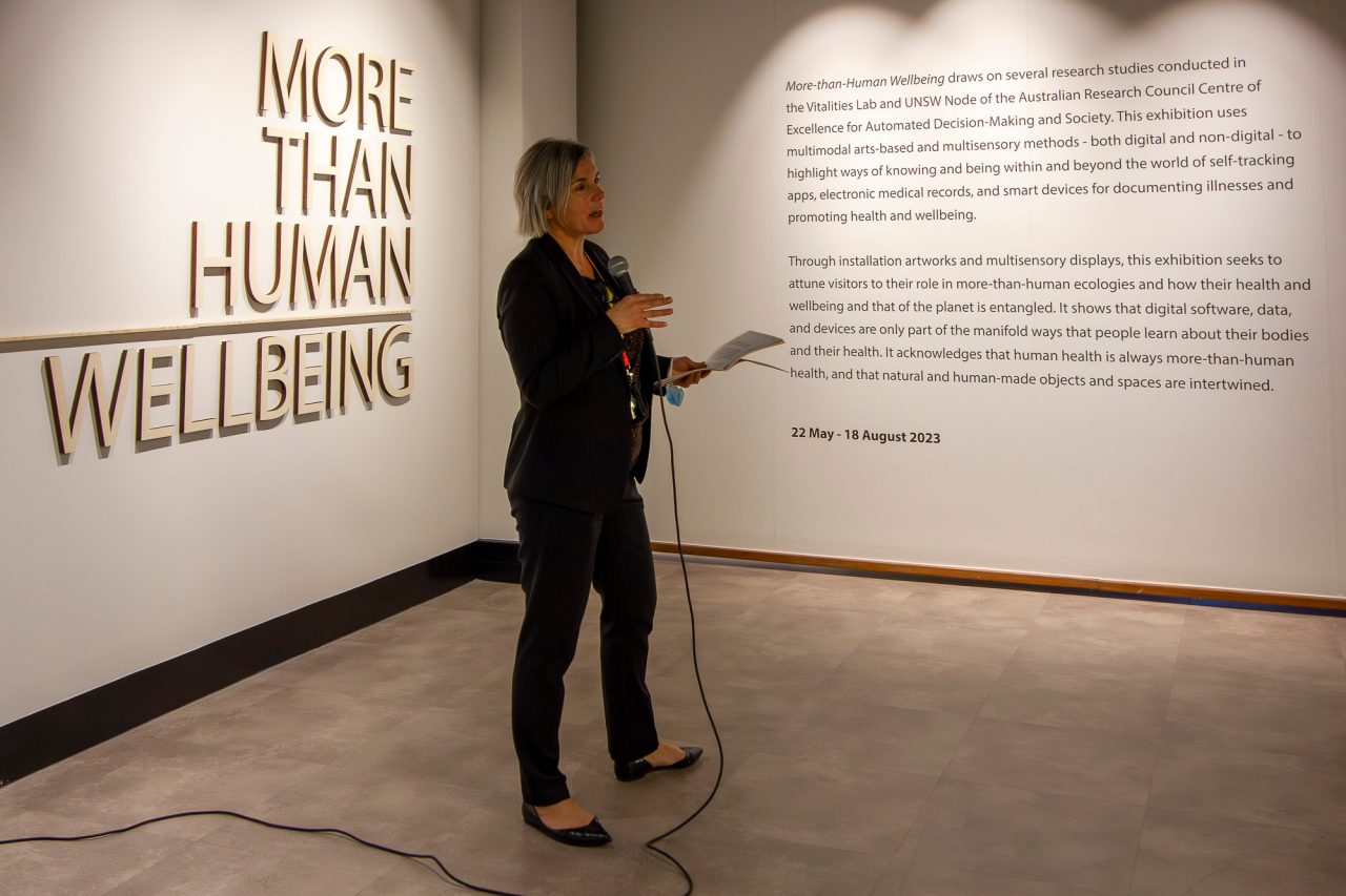 A woman in a black suit holds a microphone while giving a speech in a dimly lit room