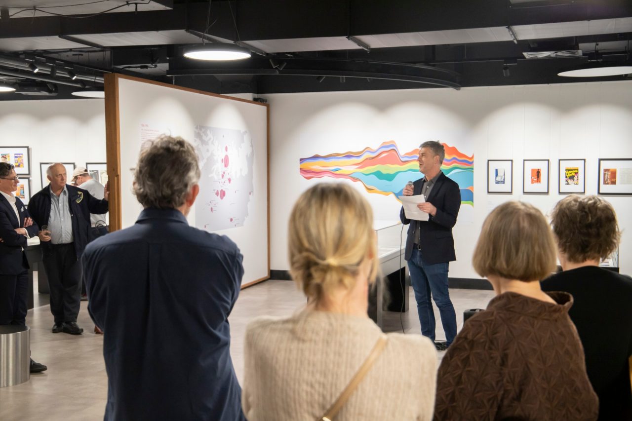 A crowd of people look on as a man in a blue blazer gives a speech while holding a microphone