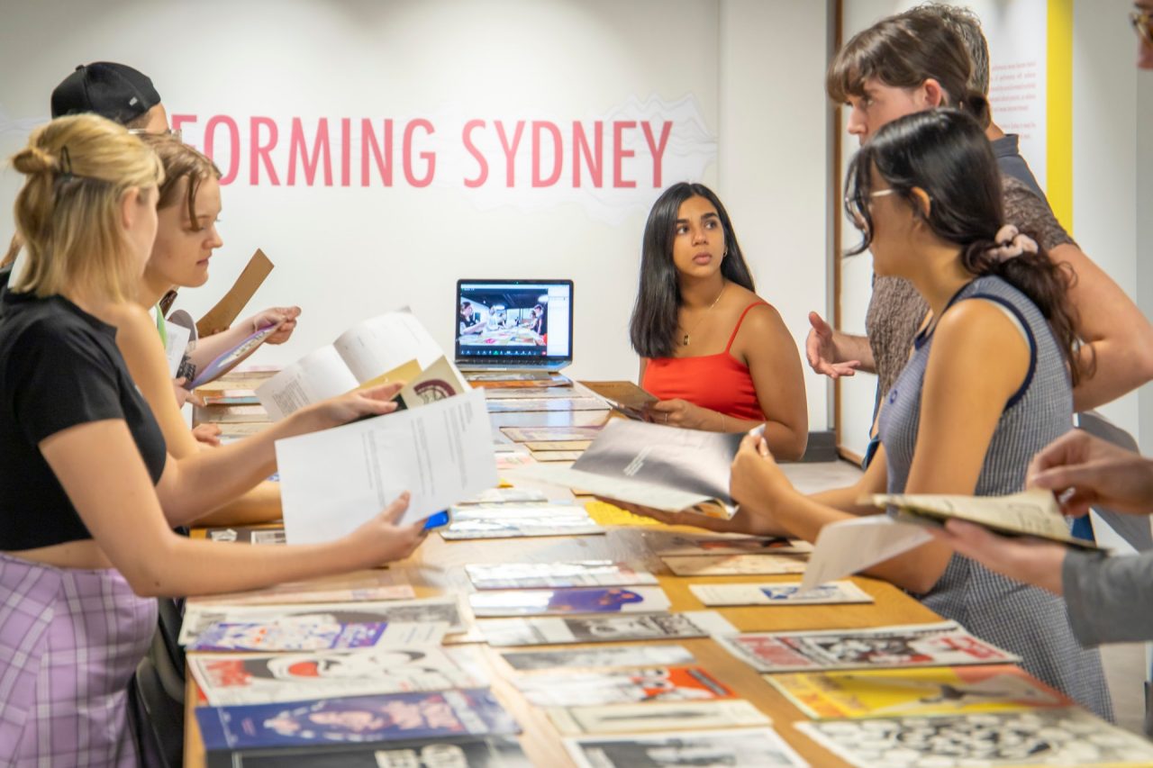 Students holding papers gather around a table covered in colourful posters. 