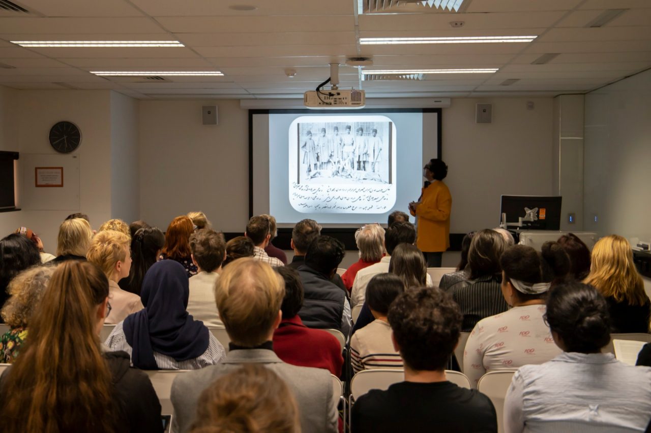 A seated crowd facing forward towards a projection listens to a man speak 