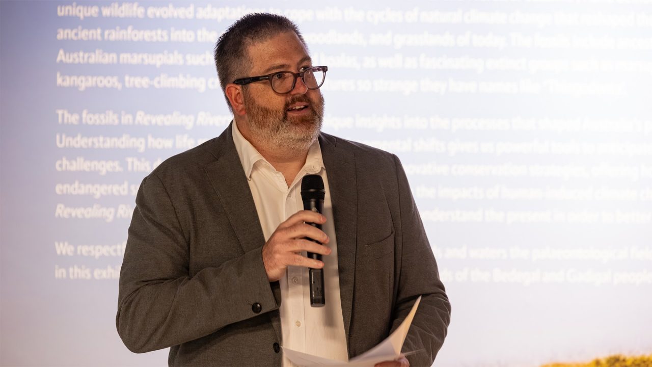 A man wearing a blazer and holding a microphone addresses an audience with a friendly smile