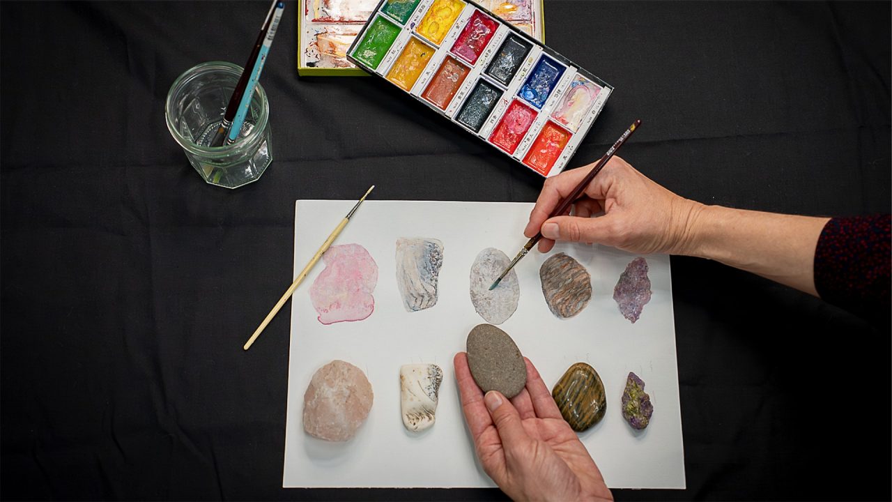 Close up of a table with watercolour paints and someone creating a colourful painting of different coloured rocks.