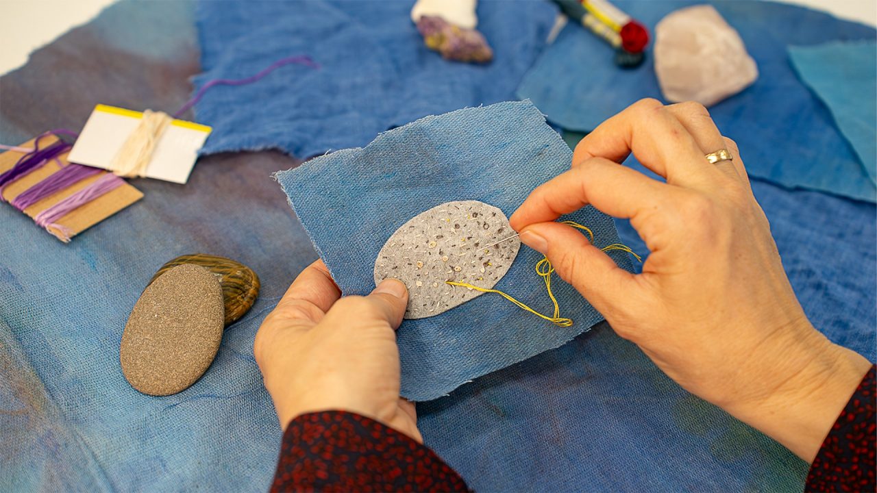 Close up of a hand embroidering on a piece of grey fabric attached to a piece of denim. The grey fabric resembles the shape of a rock.