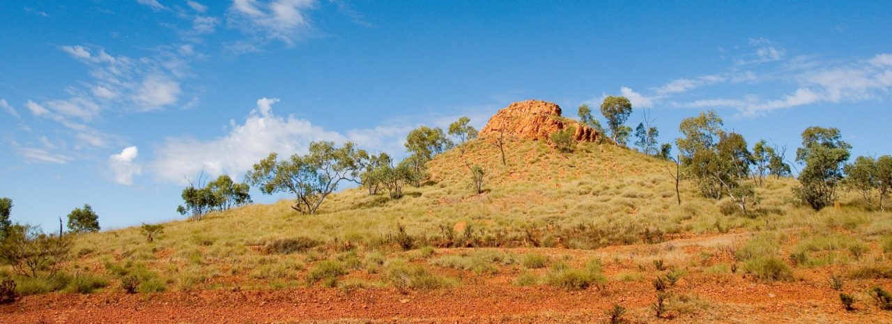 A vast, semi-arid landscape revealing orange dirt, green shrubbery, and a small hill peaking with a cluster of large rocks. 