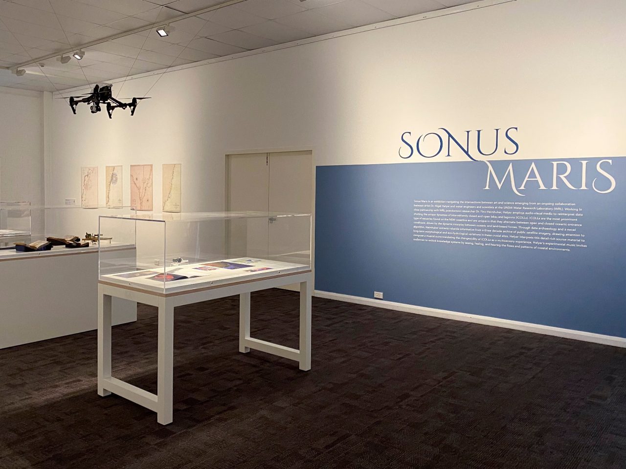A corner of the Sonus Maris exhibition with a pale-blue wall to the right with the words 'Sonus Maris' in a stylised, uppercase typeface. Several display cases are positioned in the space and a drone hangs suspended from the ceiling.