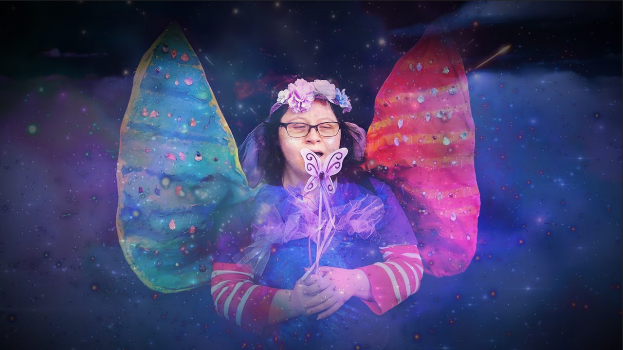 A woman with her eyes closed wearing a flower crown and green and pink fairy wings holding a butterfly wand. The background is a swirling purple mist with stars.