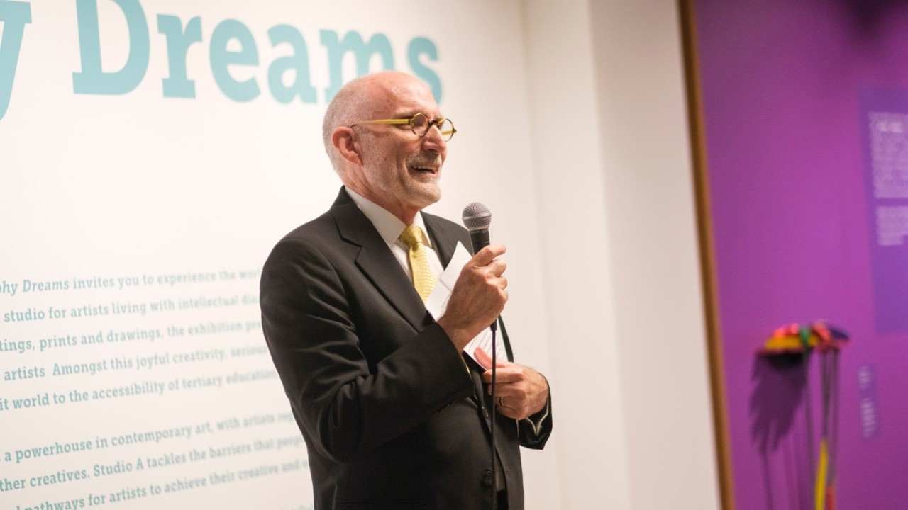 Speaker in suit holding microphone speaking to unseen crowd, standing in front of exhibition information wall.