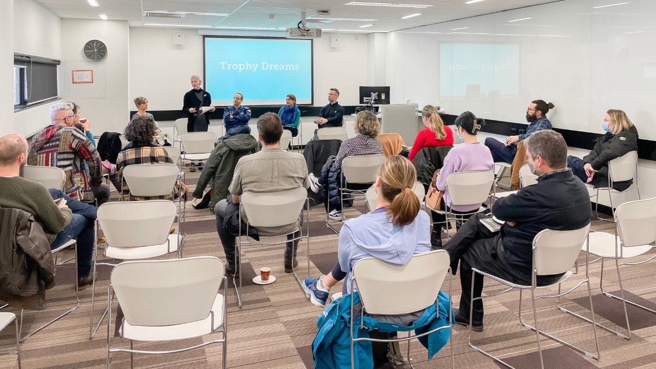 An audiences sitting in white chairs faces a panel of five seated speakers in front of a blue screen reading 'Trophy Dreams' in white letters.