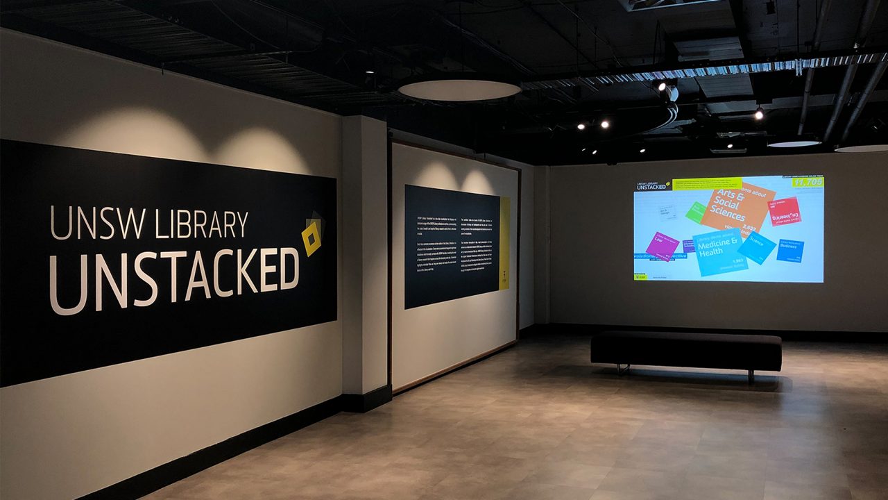 A dark gallery space with the words “UNSW Library Unstacked” displayed on a wall in large text. In the background is a video projection of a colourful data graphic.