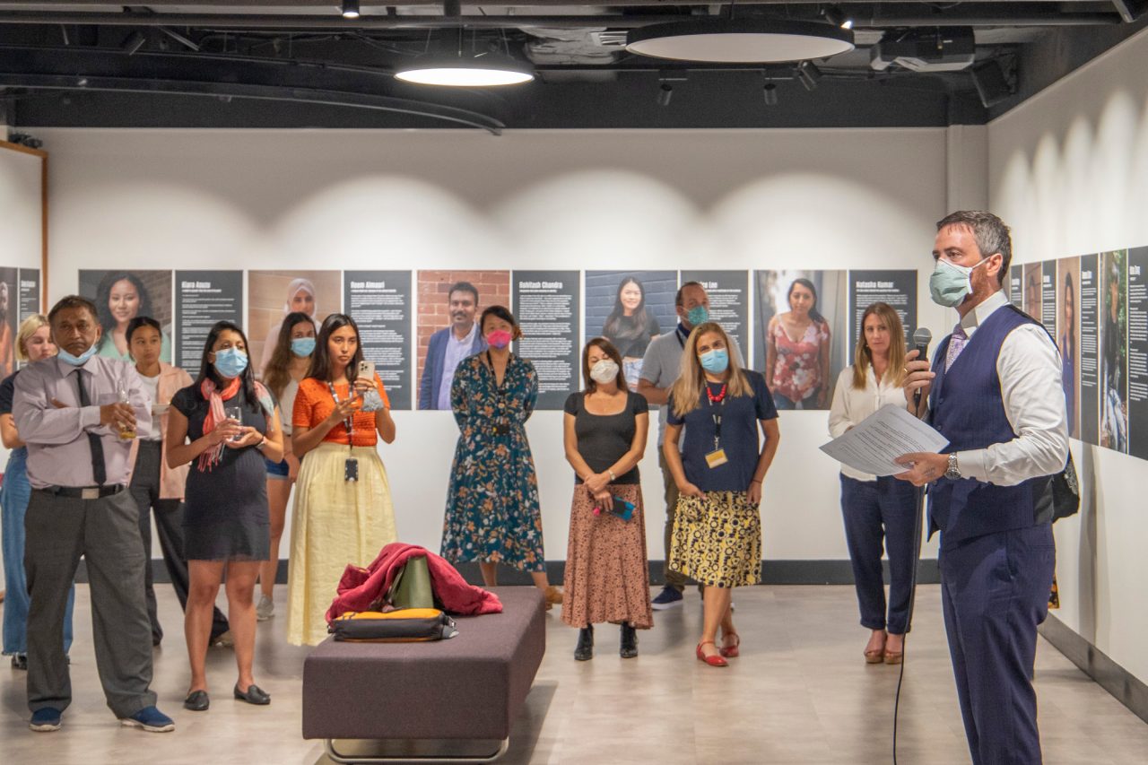 A man in a blue suit wearing a face mask is speaking to an audience at an exhibition opening