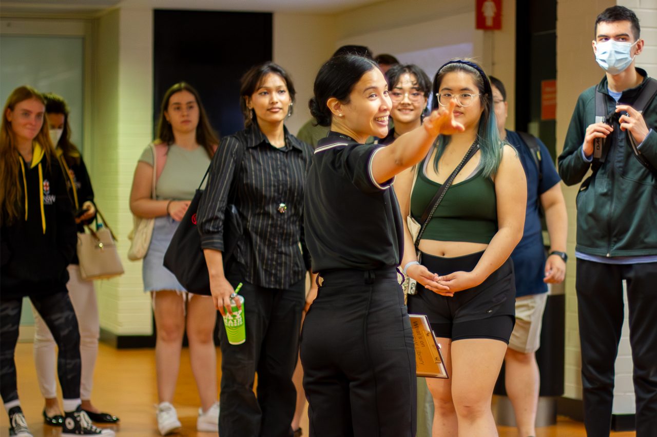 Library staff member stands in front of a group of students looking towards her. Staff member is smiling and turning to right, pointing in that direction.