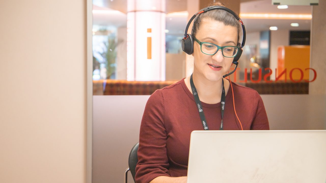 Female librarian with head set on and using laptop sits in a room with glass wall behind saying CONSULTATION in red lettering (mirror image) and orange i on white pole in distance.