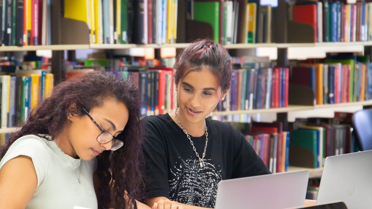 Two students sitting side by side with a laptop in front of them. They are studying and one student is talking to the other.