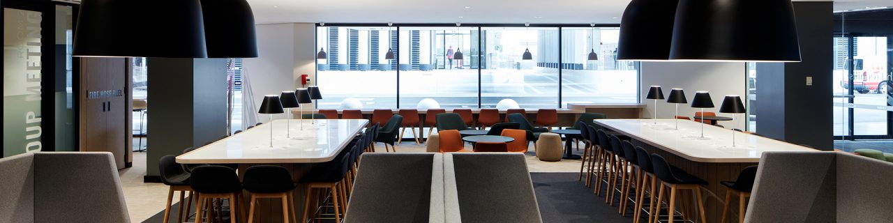 Eating area showing high benches and low seat style stools, single lounge chairs and low tables and full width bench at windows behind. Top of lounges and hanging light shades in foreground with full length windows overlooking road in background.