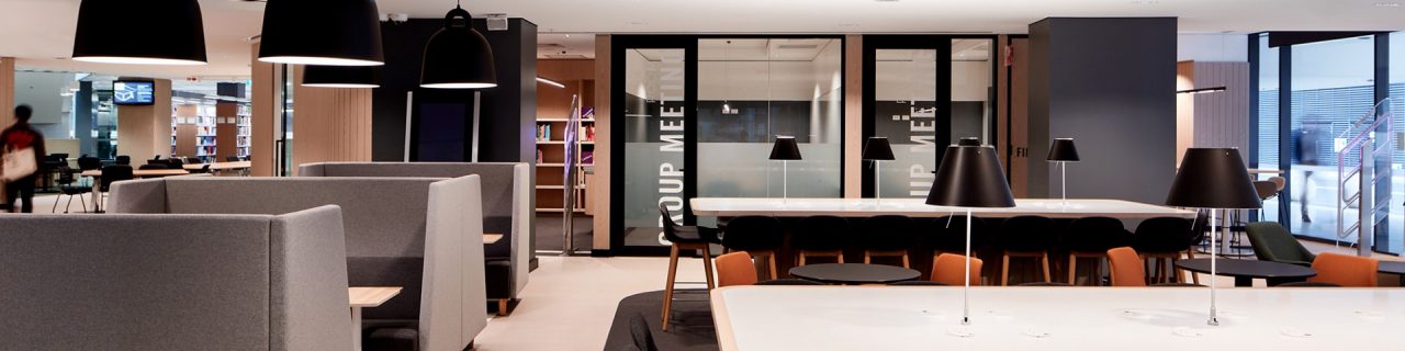 Entrance lounge view showing high benches, tables and high backed enclosed lounges with black desk lamps and hanging ceiling lights. In background are the glass fronted study rooms.
