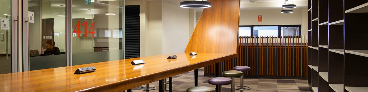 A large wooden bench desk next to glass walled group study rooms.