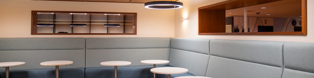 Corner of study room with light blue-grey high backed bench lounges lined along the walls and small round tables. Long rectangular alcove windows above lounges in each wall lined with wood open to areas behind.